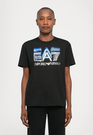 Osoba s krátkymi vlasmi, oblečená v čiernom tričku s modrou a striebornou flitrovanou nášivkou "EA7 Emporio Armani" na jednoduchom bielom pozadí.