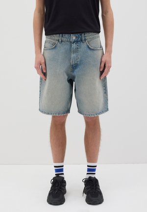 Shorts de mezclilla en un lavado claro, con corte recto, cinco bolsillos y cierre de botón. Llevados con zapatillas negras y calcetines de rayas.