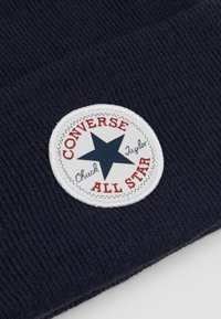 Bonnet marin en tricot ribbed, avec un patch circulaire blanc bordé de rouge, une étoile bleue et le texte "Converse All Star".