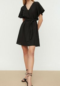Robe noire courte avec un décolleté en V, un devant à boutons et de courtes manches flottantes. Comprend une ceinture à nouer à la taille et une texture lisse. Portée avec des sandales noires.
