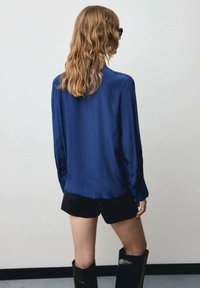 Blusa blu in satin a maniche lunghe con orlo curvo, abbinata a pantaloni corti neri e stivali neri fino al ginocchio. Capelli ondulati visibili da dietro.