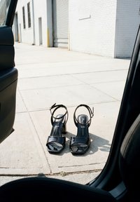 Paire de sandales à talons en cuir verni noir posées sur un trottoir ensoleillé à l'extérieur d'une porte de voiture ouverte près d'un bâtiment en briques blanches.