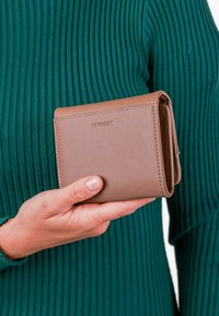 Hnědá kožená peněženka s texturovaným povrchem, s překlápěcím designem a embosovaným logem, držena proti zelenému žebrovanému svetru.