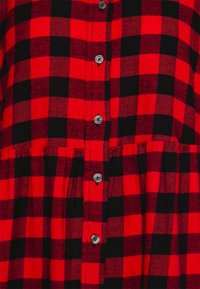 Camicia a quadri rossa e nera con chiusura frontale a bottoni, tessuto lavorato e orlo arricciato. Sei bottoni rotondi lungo la patta.