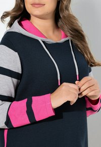 Felpa con cappuccio di colore blu navy, maniche a righe grigie e rosa, e interno del cappuccio rosa. Presenta lacci con estremità metalliche. Texture morbida.