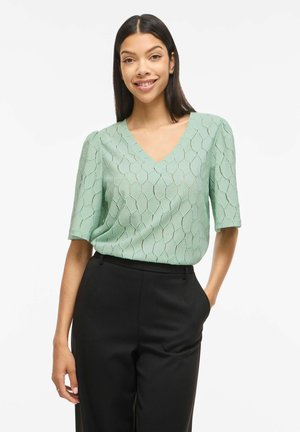 Femme souriante portant un haut vert clair texturé à col en V avec des manches aux coudes et un pantalon noir taille haute, debout avec une main dans la poche.