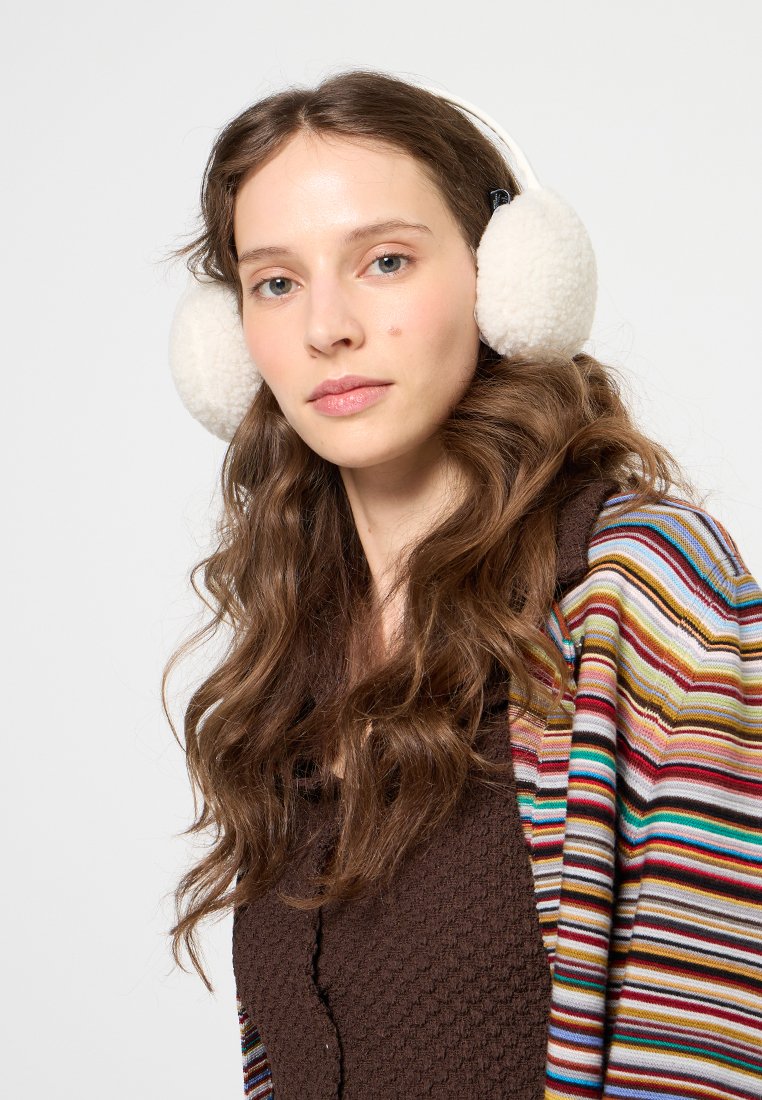 Junge Frau mit langen, welligen braunen Haaren, die weiße, flauschige Ohrenschützer, ein braunes, strukturiertes Oberteil und eine mehrfarbig gestreifte Strickjacke vor einem weißen Hintergrund trägt.