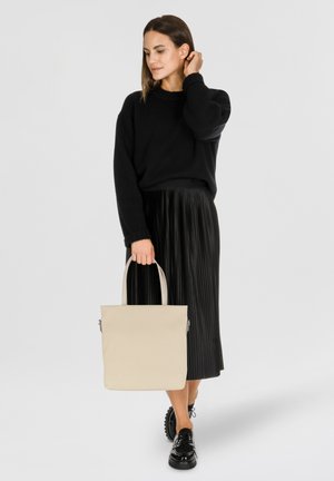 Kobieta w czarnym swetrze i plisowanej spódnicy, trzymająca beżową torbę typu tote, patrząca w bok, jedną ręką dotykająca włosów.