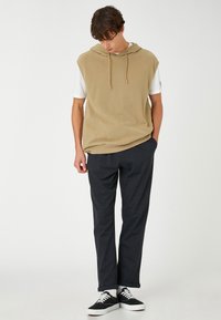 Beige mouwloze hoodie met een trekkoord, gedragen over een wit shirt, in combinatie met donkere broeken en zwarte sneakers. Soepele stoffen en een relaxte pasvorm.