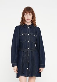 Robe en denim à manches longues, col et devant boutonné. Dotée de deux poches poitrine, d'une taille ceinturée et d'une couleur bleu foncé avec des coutures discrètes.