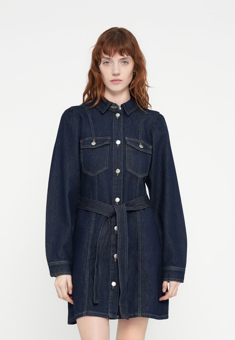 Robe en denim à manches longues, col et devant boutonné. Dotée de deux poches poitrine, d'une taille ceinturée et d'une couleur bleu foncé avec des coutures discrètes.