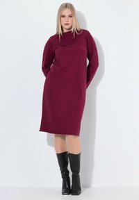 Abito bordeaux al ginocchio con scollatura a barca, maniche lunghe e tasche laterali, abbinato a stivali neri alti fino al ginocchio e una texture morbida.