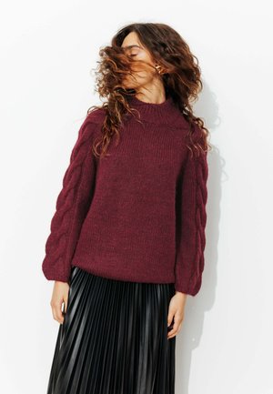 Pull en maille bourgogne avec un col montant et des manches en tricot torsadé, associé à une jupe plissée en simili cuir noir. Texture lisse et coupe décontractée.