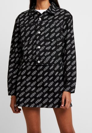 Black buttoned jacket dress with long sleeves and white "CK50" logos patterned all over, worn by a person with bare legs.