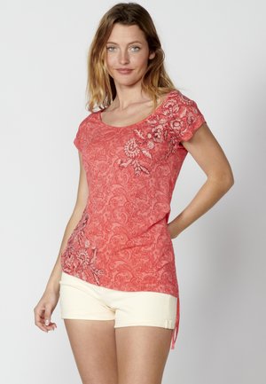 Haut à motifs corail avec des manches courtes, encolure dégagée, et broderie florale d'un côté. Associé à un short beige clair.