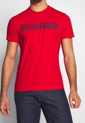 Homme portant un T-shirt à manches courtes rouge vif avec l'inscription « NAPAPIJRI geographic » en noir sur la poitrine, associé à un jean bleu foncé.