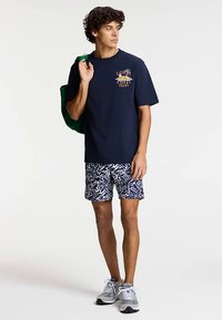 Tengerészkék pamut póló grafikai mintával, kék-fehér mintás rövidnadrággal párosítva. A modell zöld táskát tart, szürke sportcipőt visel.