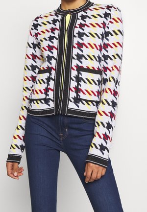 Femme portant un cardigan à motif pied-de-poule multicolore avec des bordures noires et des poches avant, assorti à un jean bleu foncé ajusté.