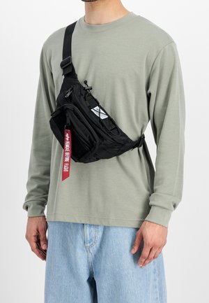 Homme portant une chemise à manches longues vert clair et un jean bleu clair avec un sac bandoulière noir doté d'une étiquette rouge "Remove Before Flight".