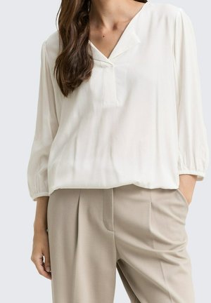 Femme portant un chemisier blanc ample à manches trois-quarts et un pantalon beige taille haute, avec une main dans la poche.