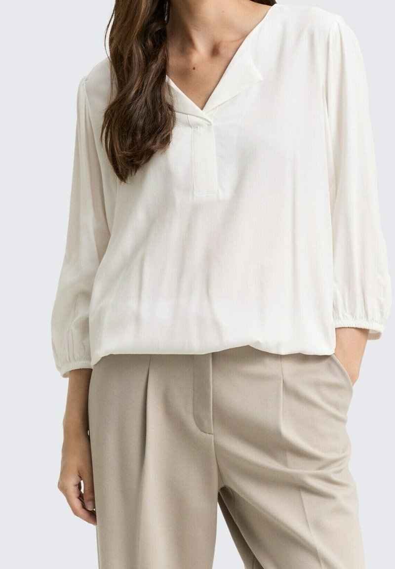 Femme portant un chemisier blanc ample à manches trois-quarts et un pantalon beige taille haute, avec une main dans la poche.