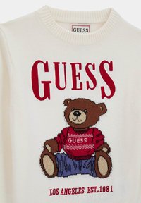 Pull en tricot crème avec un ours en cartoon portant un pull rouge et un jean bleu. Logo "GUESS" en rouge vif au-dessus de l'ours.