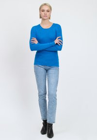 Maglione a maniche lunghe blu a coste con scollo rotondo, abbinato a jeans azzurri chiari e stivaletti neri alla caviglia. Sfondo semplice.