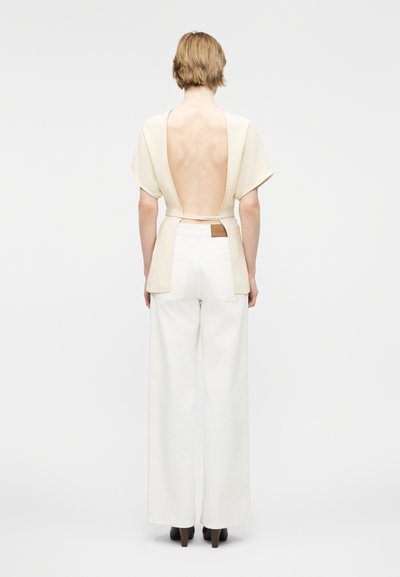 Personne aux cheveux blonds courts portant un haut beige dos nu et un pantalon blanc large à taille haute, debout devant un fond uni.