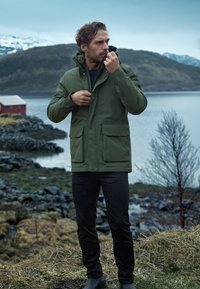 Oliivivihreä vedenpitävä takki, jossa on korkea kaulus, etutaskut ja vetoketju, yhdistetty mustiin housuihin ja saappaisiin kiviseen maisemaan.