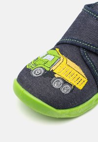 Blå lærredssko med grøn gummisål, der har et gult og grønt broderet dumperdesign på tåområdet. Justerbar remlukning.