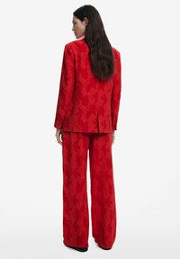 Rode op maat gemaakte suit met een gestructureerd loofpatroon, met een aangesloten blazer en wijde broekspijpen. De achterkant toont knoopdetails en strakke lijnen.