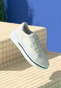 Weiße Low-Top-Sneaker mit Schnürsenkeln und einem schwarzen Streifen auf der Sohle, die auf beigen Fliesenstufen vor einem blauen Wandhintergrund platziert sind.