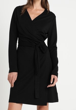 Frau trägt ein schwarzes, langärmeliges Wickelkleid, das an der Taille gebunden ist, mit V-Ausschnitt und knielangem Saum.