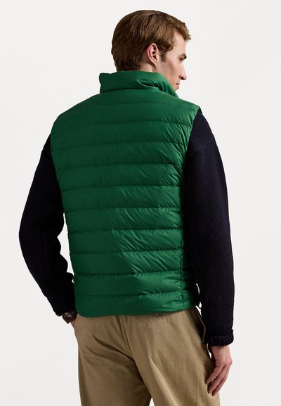 Green puffer vest with a high collar, featuring horizontal quilted sections, worn over a dark navy sweater. Includes visible cuff details.