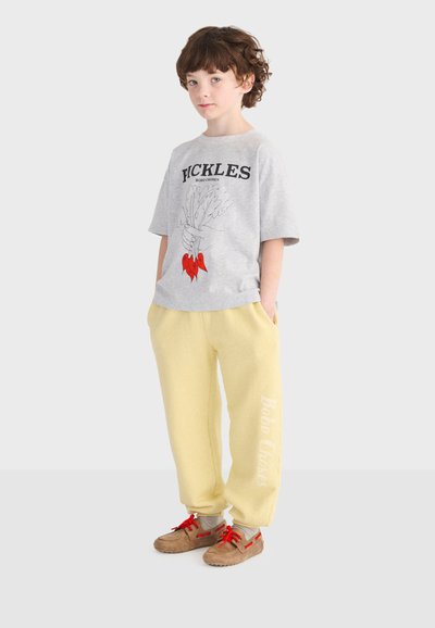 Enfant aux cheveux bouclés portant un T-shirt gris avec un motif de légumes, un pantalon de survêtement jaune et des chaussures marron avec des lacets rouges, debout les mains dans les poches.