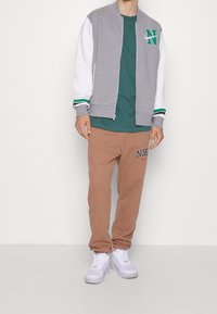 Giacca bomber grigia e bianca con dettagli verdi, abbinata a pantaloni felpati marroni con il logo rosso "NIKE". Sneakers bianche completano l'outfit.