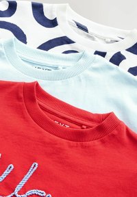Tres camisetas de cuello redondo: blanca con un patrón azul marino, azul claro y roja con bordado azul. Todas cuentan con cuellos acanalados y están hechas de algodón.