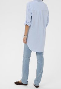 Chemise bleu clair à manches longues avec poignets retroussés, empiècement arrière boutonné et ourlet incurvé, associée à un pantalon en denim clair et des ballerines marron.