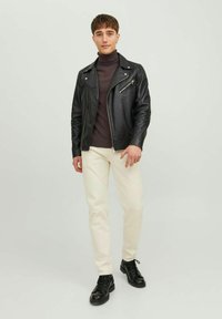 Giacca da moto in pelle nera sopra un dolcevita scuro, pantaloni color crema e stivali neri con lacci. Design semplice con dettagli metallici della cerniera.