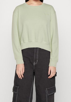 Kvinde iført lysegrøn cropped sweatshirt og sorte cargobukser med kontrasterende hvid syning, stående mod hvid baggrund.