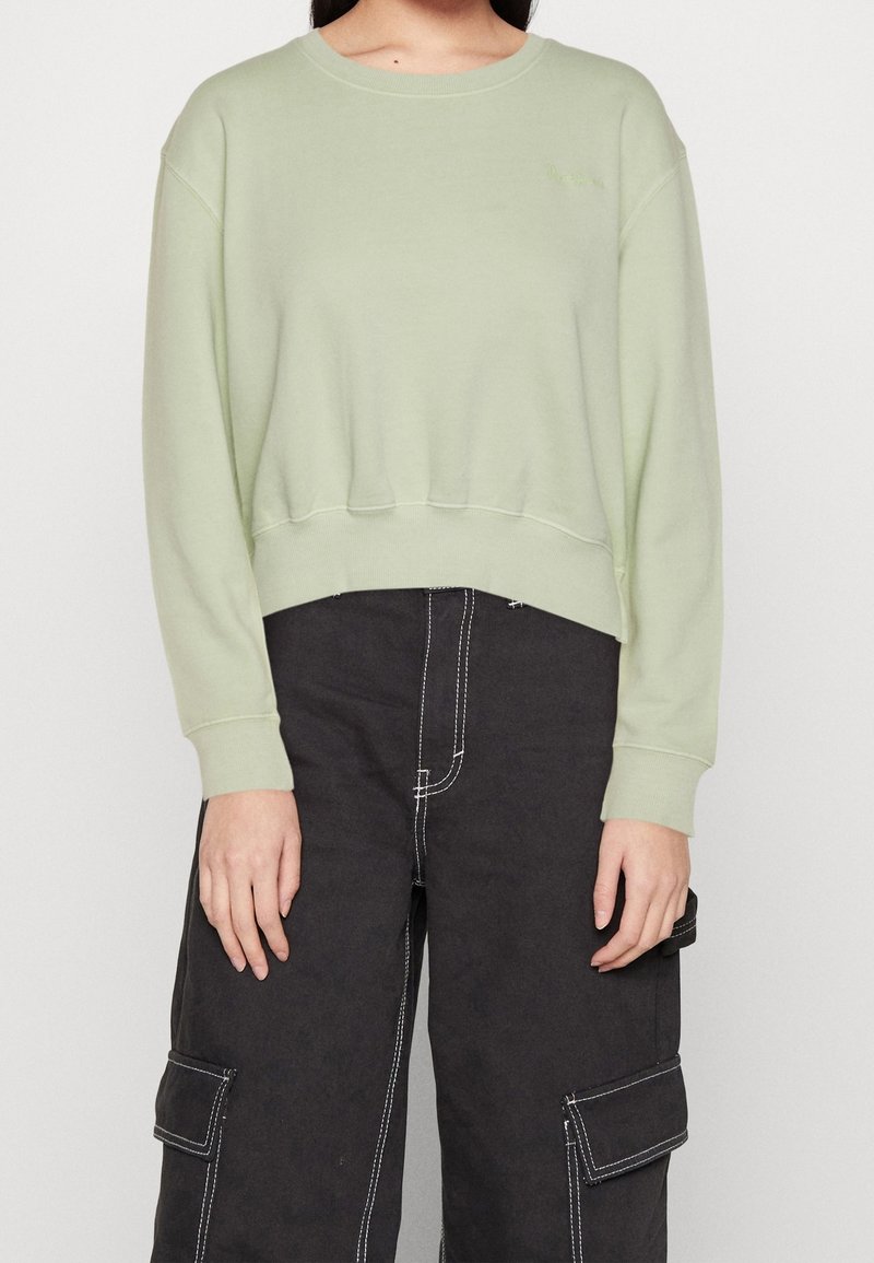 Femme portant un sweat court vert clair et un pantalon cargo noir avec des coutures blanches contrastantes, debout devant un fond blanc.