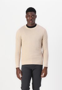 Beige gebreide trui met een gestructureerd patroon, ronde hals en geribde boorden en zoom. Draagend met zwarte broeken met een getailleerd silhouet.
