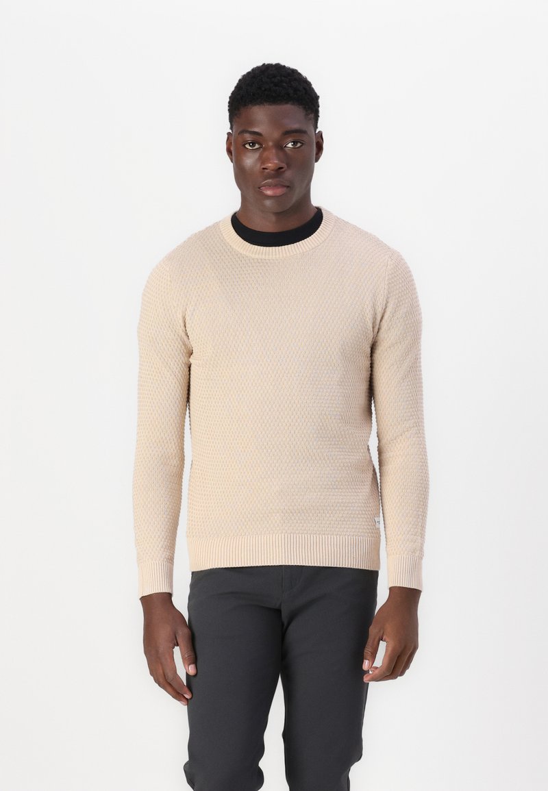 Beige gebreide trui met een gestructureerd patroon, ronde hals en geribde boorden en zoom. Draagend met zwarte broeken met een getailleerd silhouet.