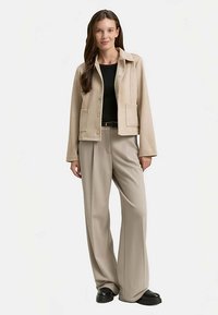 Beige cropped jack met kraag, twee voorzakken, gecombineerd met wijde beige broek en zwarte schoenen, op een neutrale achtergrond.