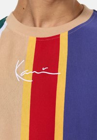 Camiseta de algodón multicolor con rayas verticales en beige, rojo, amarillo y azul, con un logo bordado en blanco en el centro.