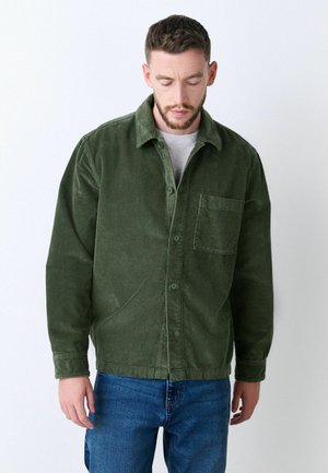 Green corduroy shirt with a button-up front, a chest pocket, and a classic collar, paired with blue jeans. Textured and durable fabric.