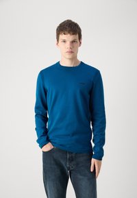 Cămașă albastră din bumbac cu mânecă lungă, cu accente ribbed pe umeri, decolteu rond și un mic logo negru pe piept. Asortată cu blugi de denim închis.