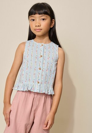 Fată tânără cu păr lung și întunecat poartă o bluză fără mâneci, cu nasturi, cu model floral și tiv cu volane, precum și pantaloni roz lejeri, pe un fundal simplu.