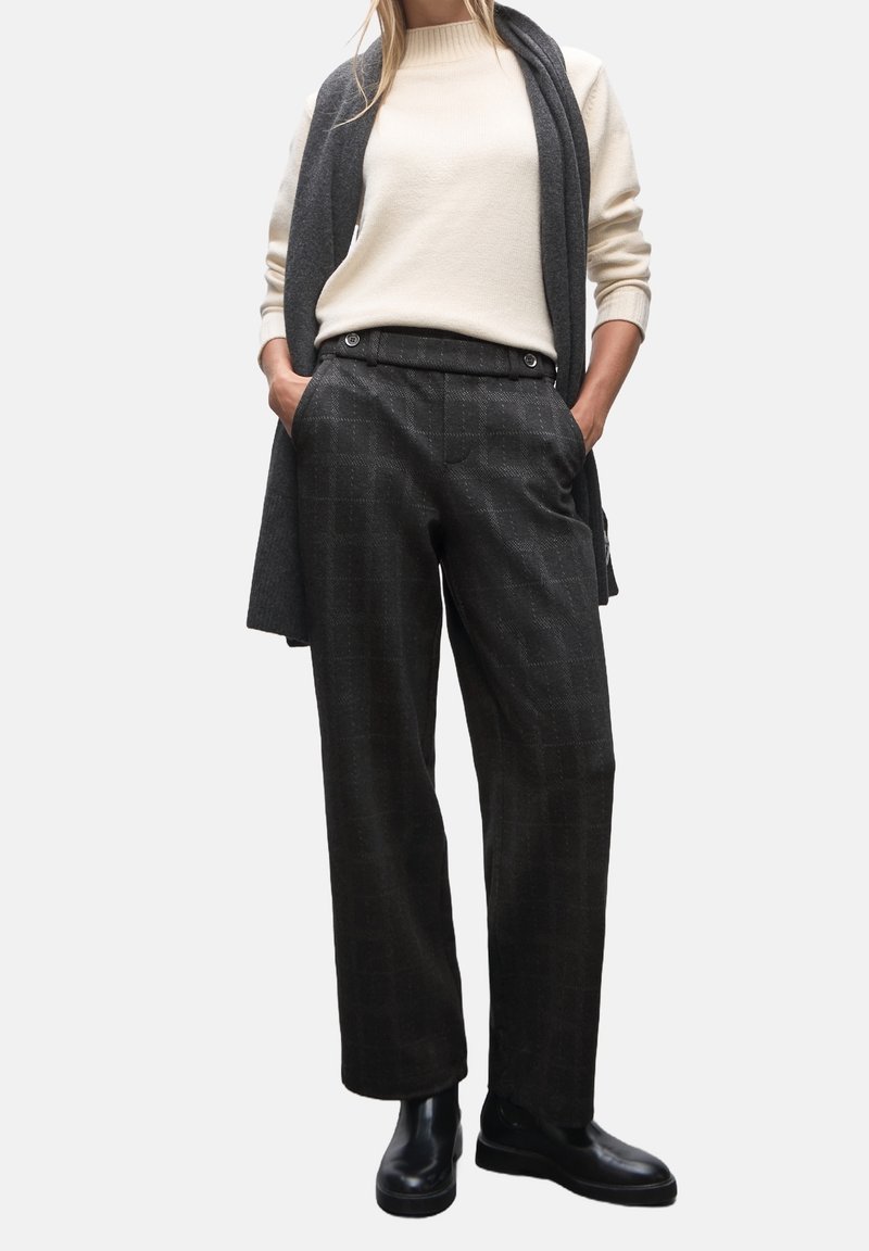 Femme portant un pull crème, un pantalon large à carreaux foncé, des bottines noires et une écharpe sombre drapée sur les épaules, les mains dans les poches.