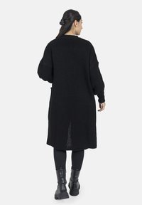 Langer schwarzer Strickcardigan mit übergroßen Ärmeln, versehen mit einem Schlitz im Rücken für Belüftung. Getragen über schwarzen Leggings und klobigen Stiefeln.